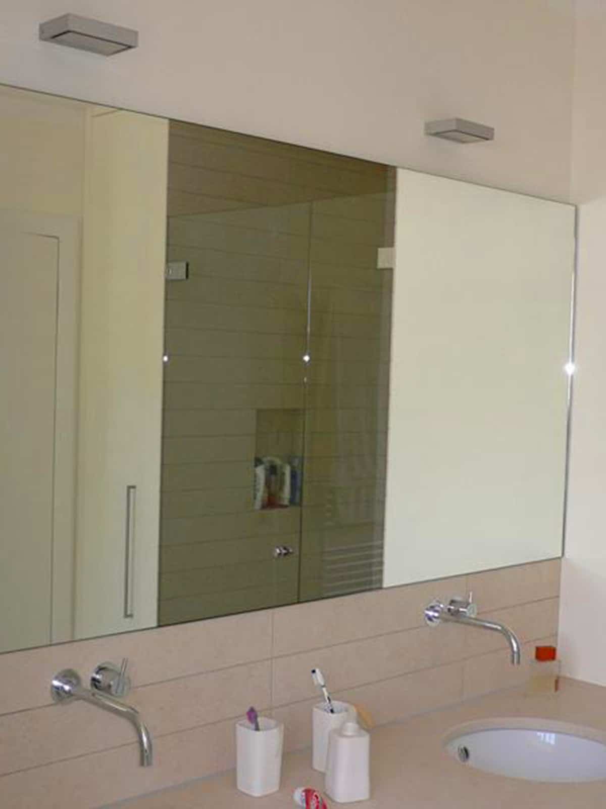 Modernes Badezimmer mit großem Wandspiegel, wandmontierten Armaturen, Waschbecken und Duschbereich im Hintergrund.