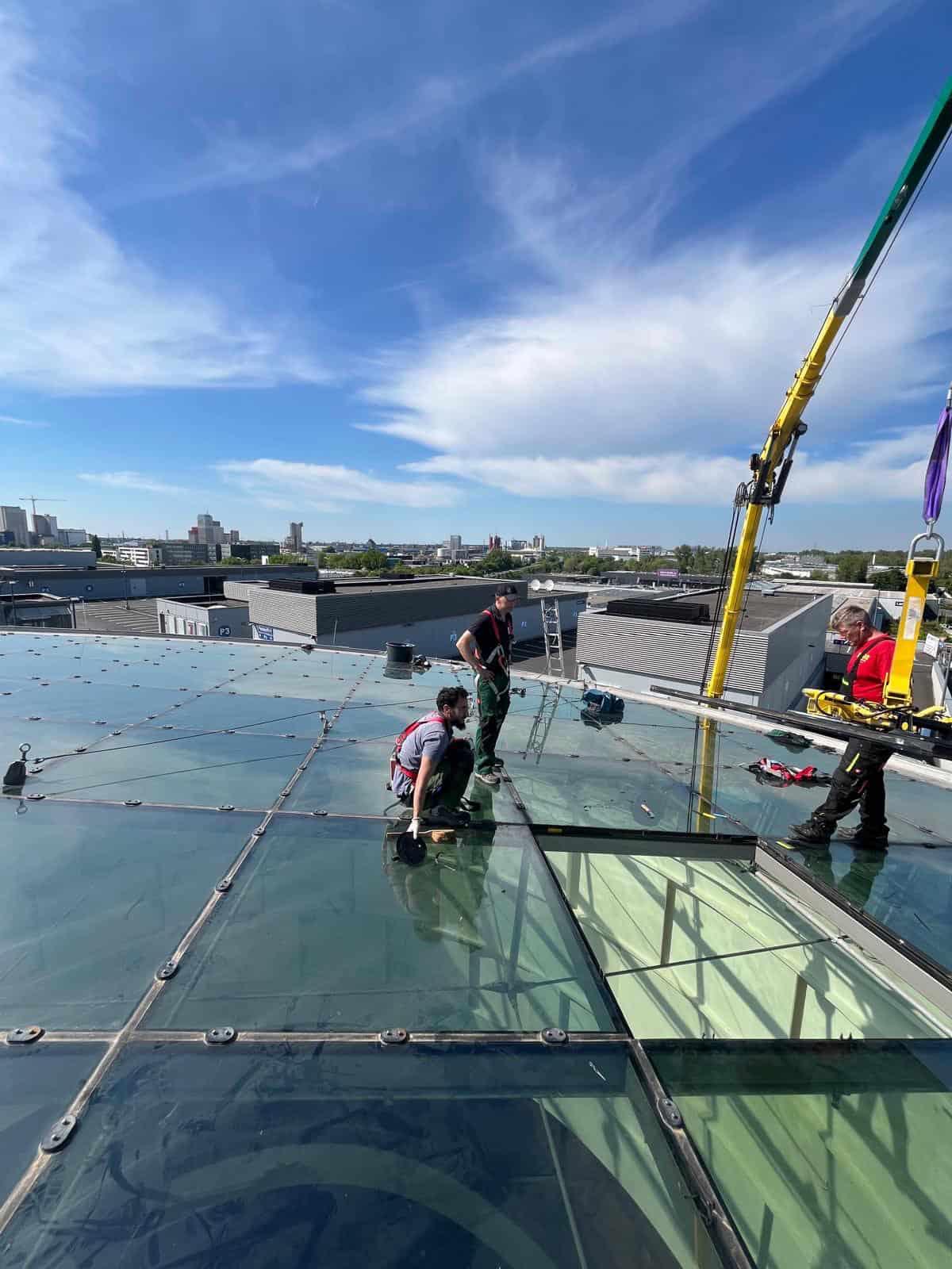 Drei Dachdecker installieren Glasplatten auf einem Flachdach mit Kranunterstützung über einer städtischen Skyline.